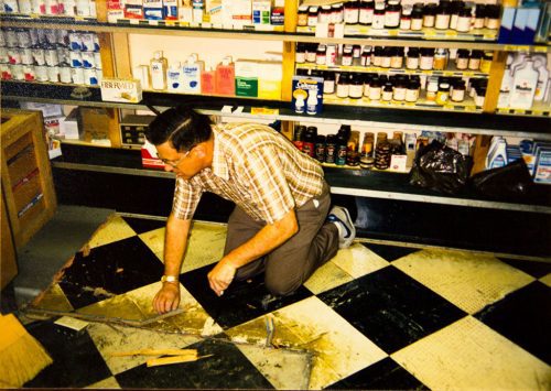 An image of Karl Hanke on his hands and knees, surrounded by shelves of products, making repairs to the checkered tile floor
